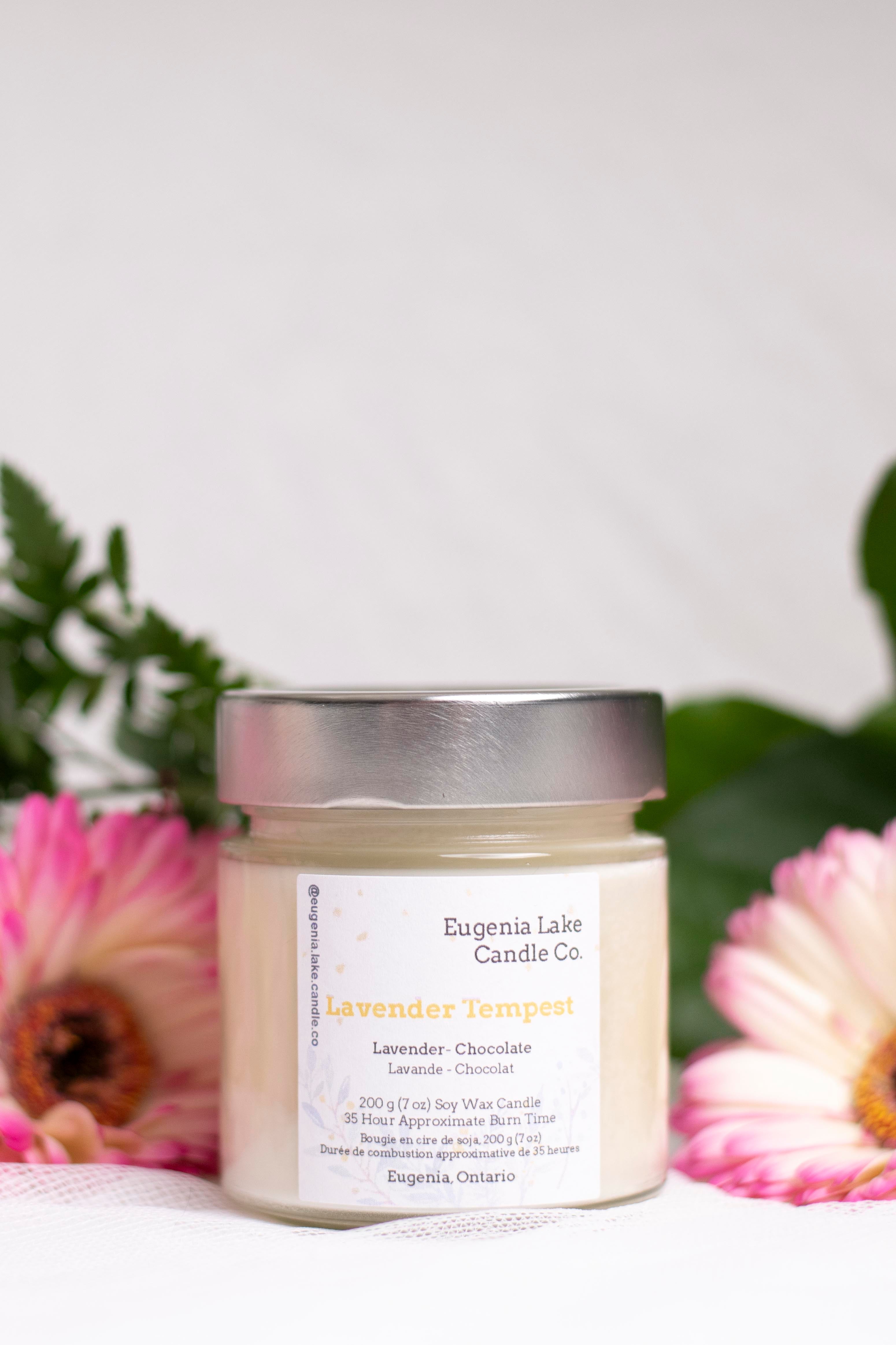A jar candle from Eugenia Lake Candle Co. labeled 'Lavender Tempest' sits on a white surface with pink flowers and green leaves blurred in the background.