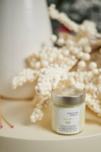 A candle in a clear jar with a silver lid and a white label on a white tray with a white vase and some matches to the side. 